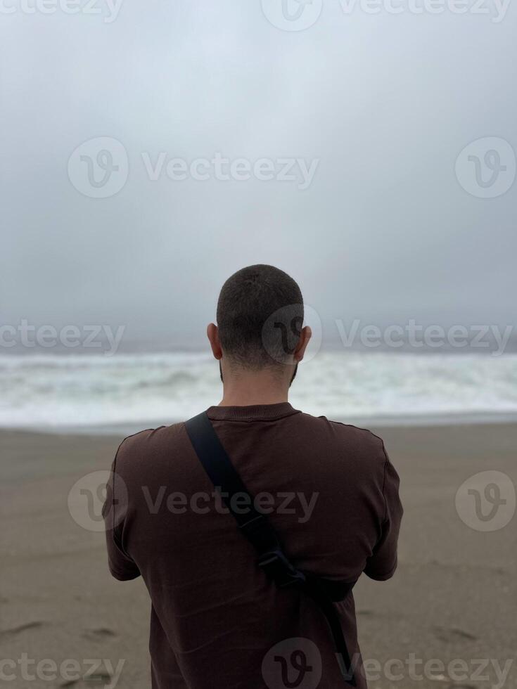 Young guy, tourist, looking at the ocean in cloudy weather, vertical photo