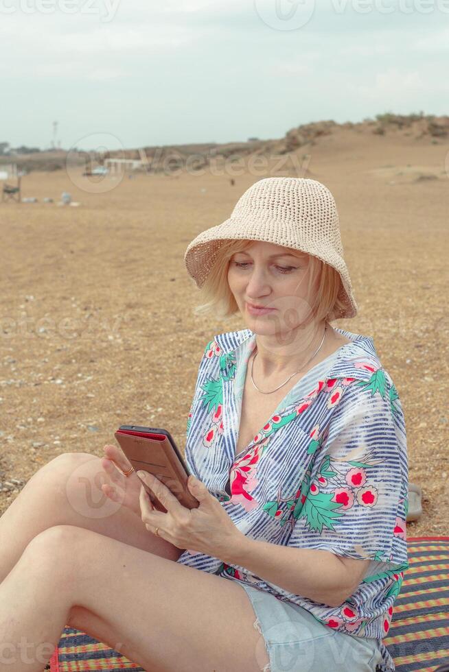 A woman in a sun hat and colorful blouse sits on a beach, holding a phone and looking at the camera under an overcast sky. photo