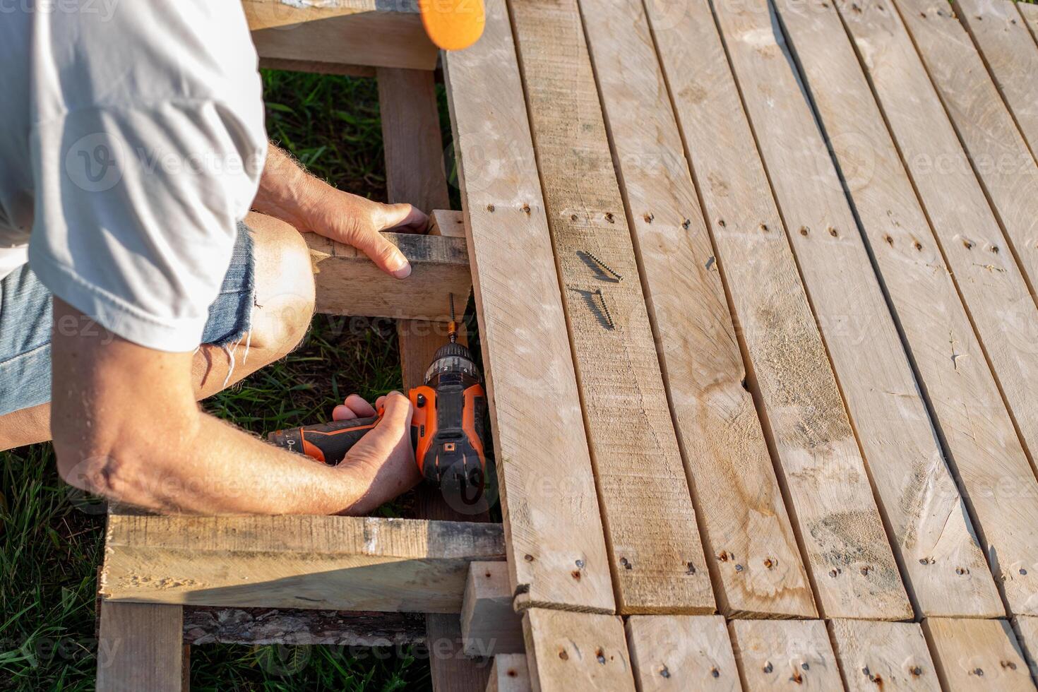 A man assembles a podium made of wooden pallets outdoors on the grass using a drill. Making your own garden furniture photo