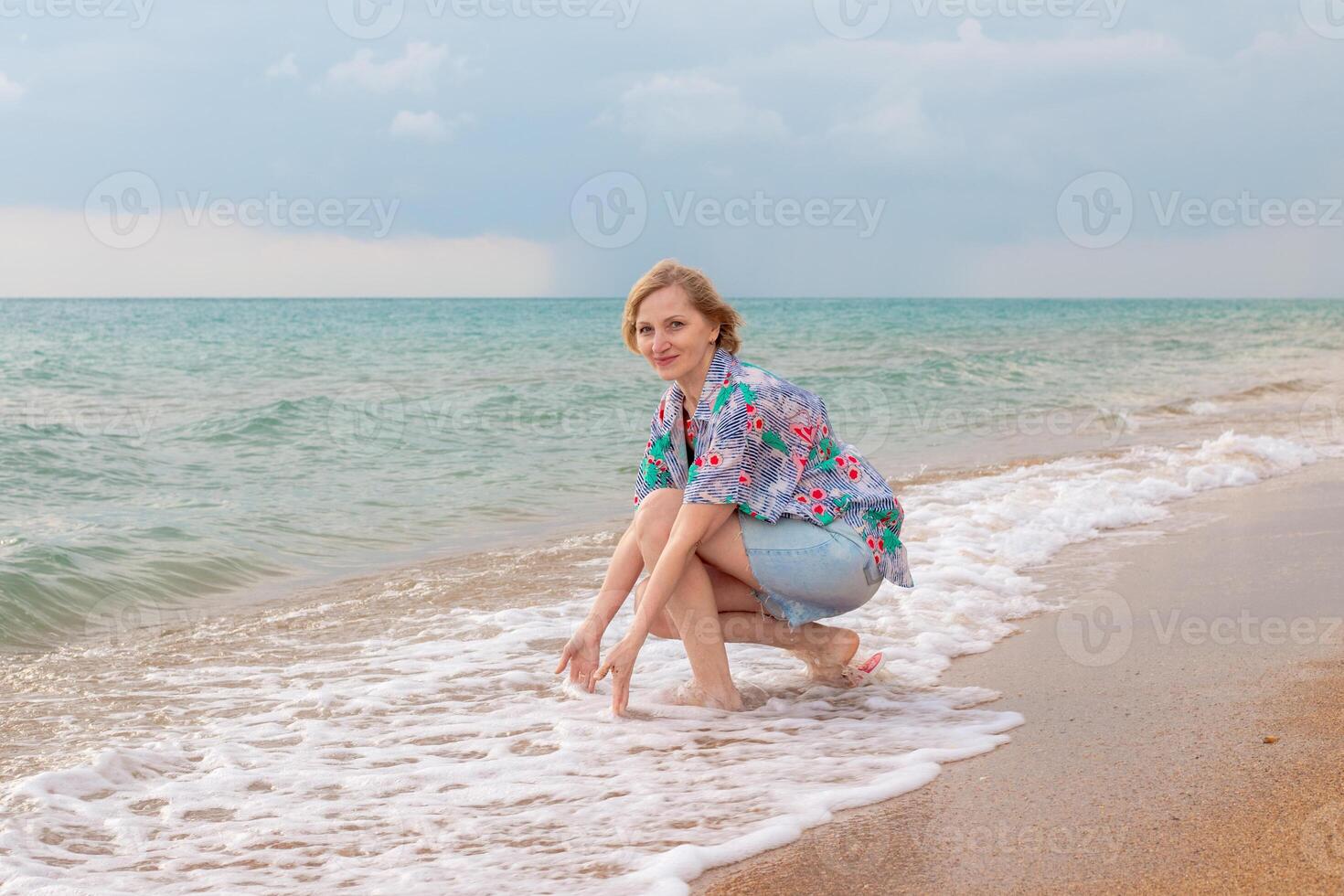 Woman in floral shirt and denim shorts squatting playing in shallow waves on sandy beach. Summer vacation at sea photo