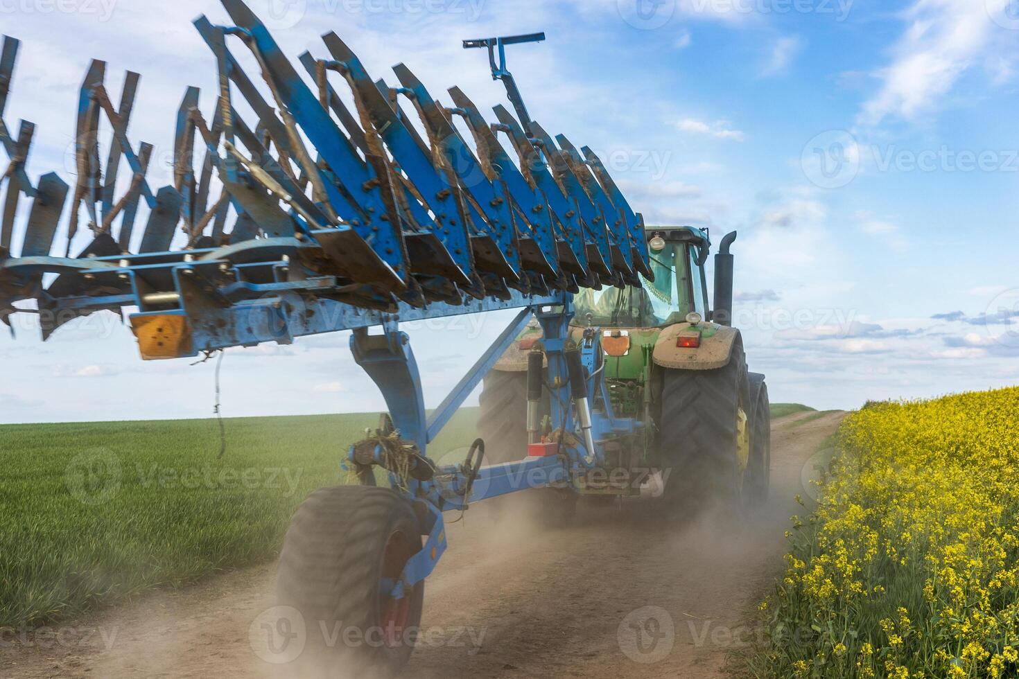 A tractor with a plow is working a large field, creating dust as it moves along a dirt path beside vibrant yellow flowers under a blue sky photo