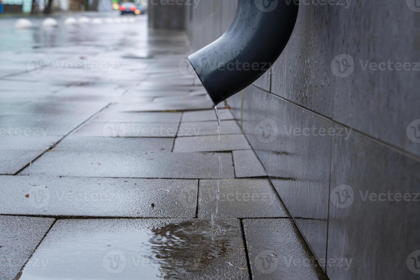 Water drips from a gutter onto rain-soaked pavement, reflecting the gray sky in a quiet city street during a rainy day photo