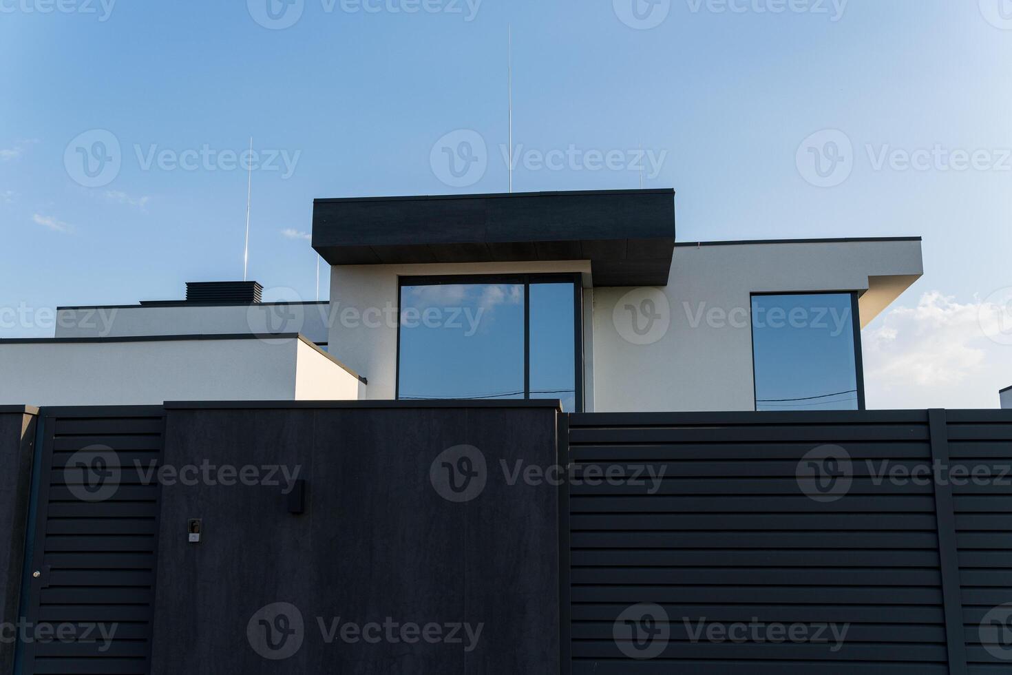 A contemporary house features minimalistic lines and large glass windows, surrounded by a modern fence under a blue sky photo