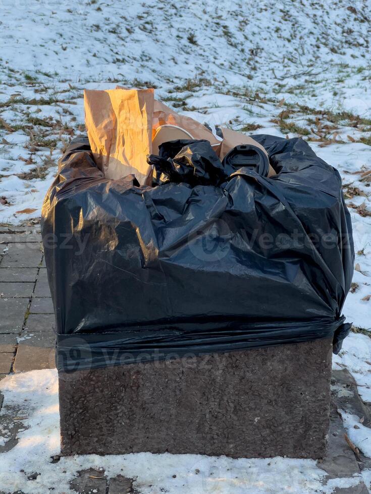 A black trash bag filled with garbage sits atop a stone pedestal near a snowy walkway in a park during late afternoon photo