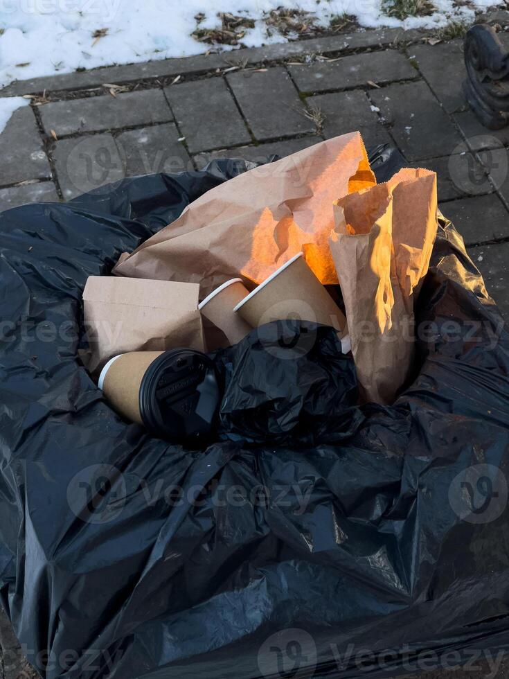 A trash bin is overflowing with various discarded food containers, paper wrappers, and bags, highlighting urban waste in wintertime photo