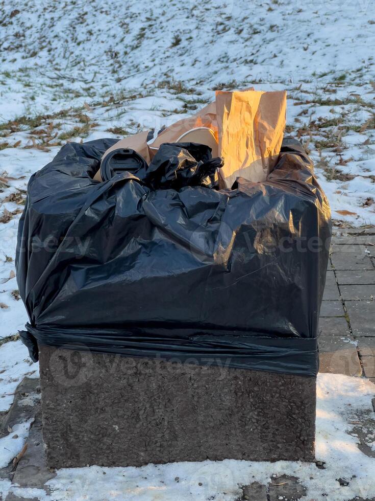 A black trash bag overflowing with waste sits on a concrete base, surrounded by patches of snow in a public area during winter photo