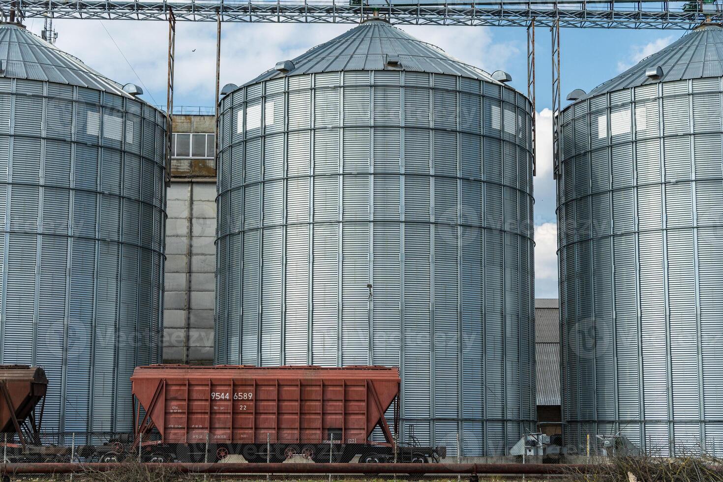 Tall metal silos store grain at an agricultural site, with a freight train wagon parked nearby under a blue sky with clouds photo