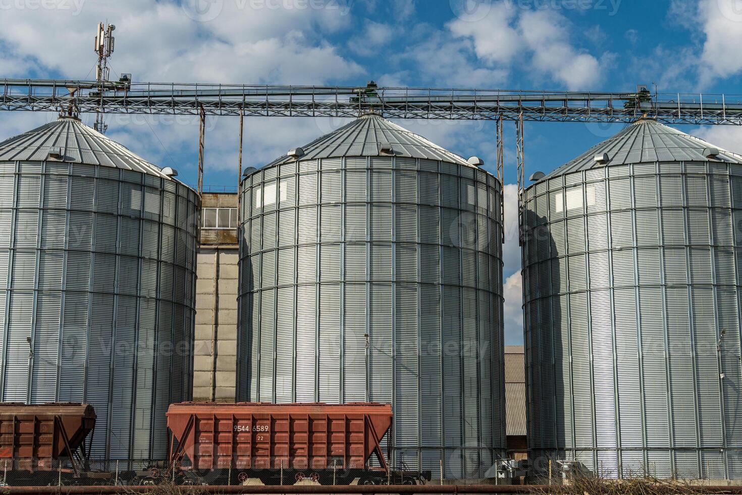 Three tall metal silos stand beside railway cars, showcasing agricultural storage infrastructure under a bright sky photo