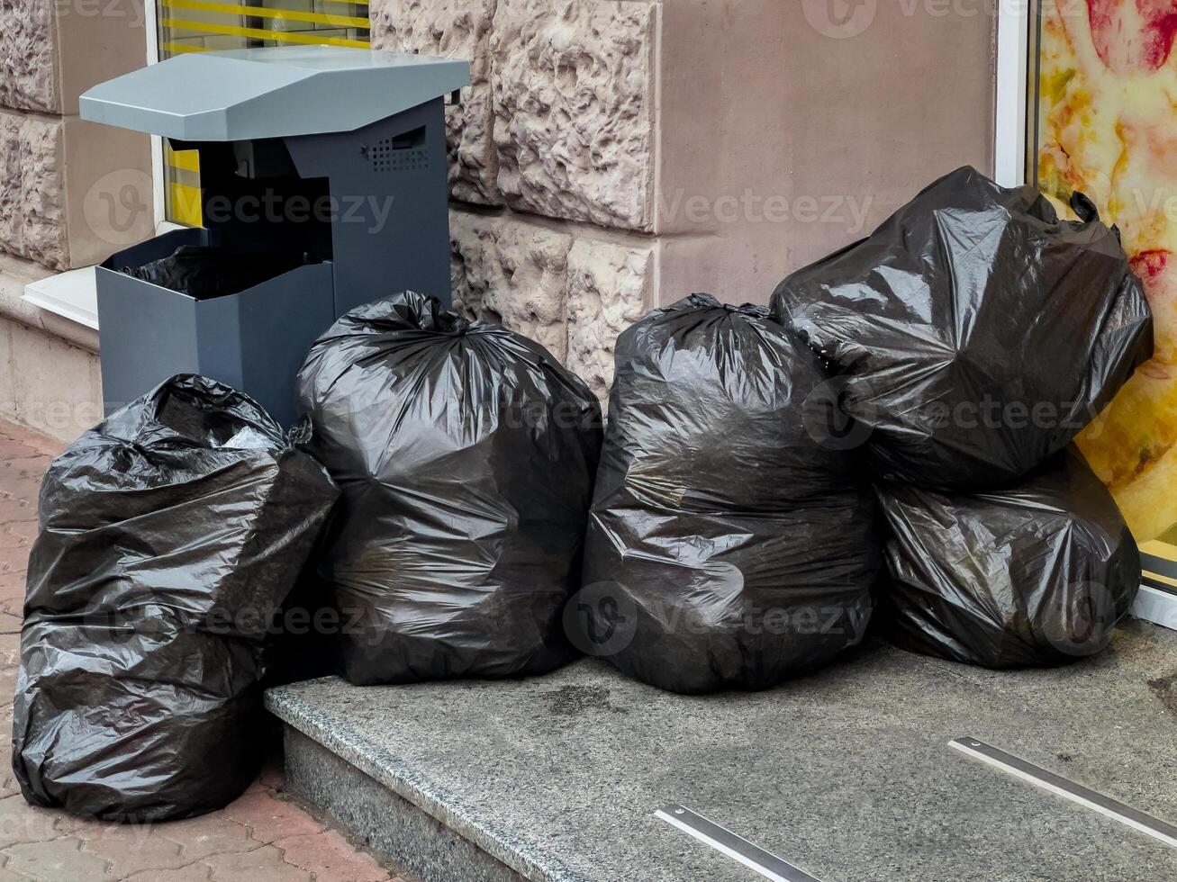 Black garbage bags are stacked next to a public trash bin outside a shop on a busy city sidewalk during the afternoon photo