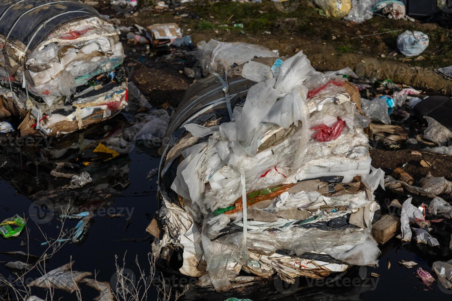 Large piles of compressed waste materials are seen near a polluted water source in an urban setting, highlighting environmental concerns photo