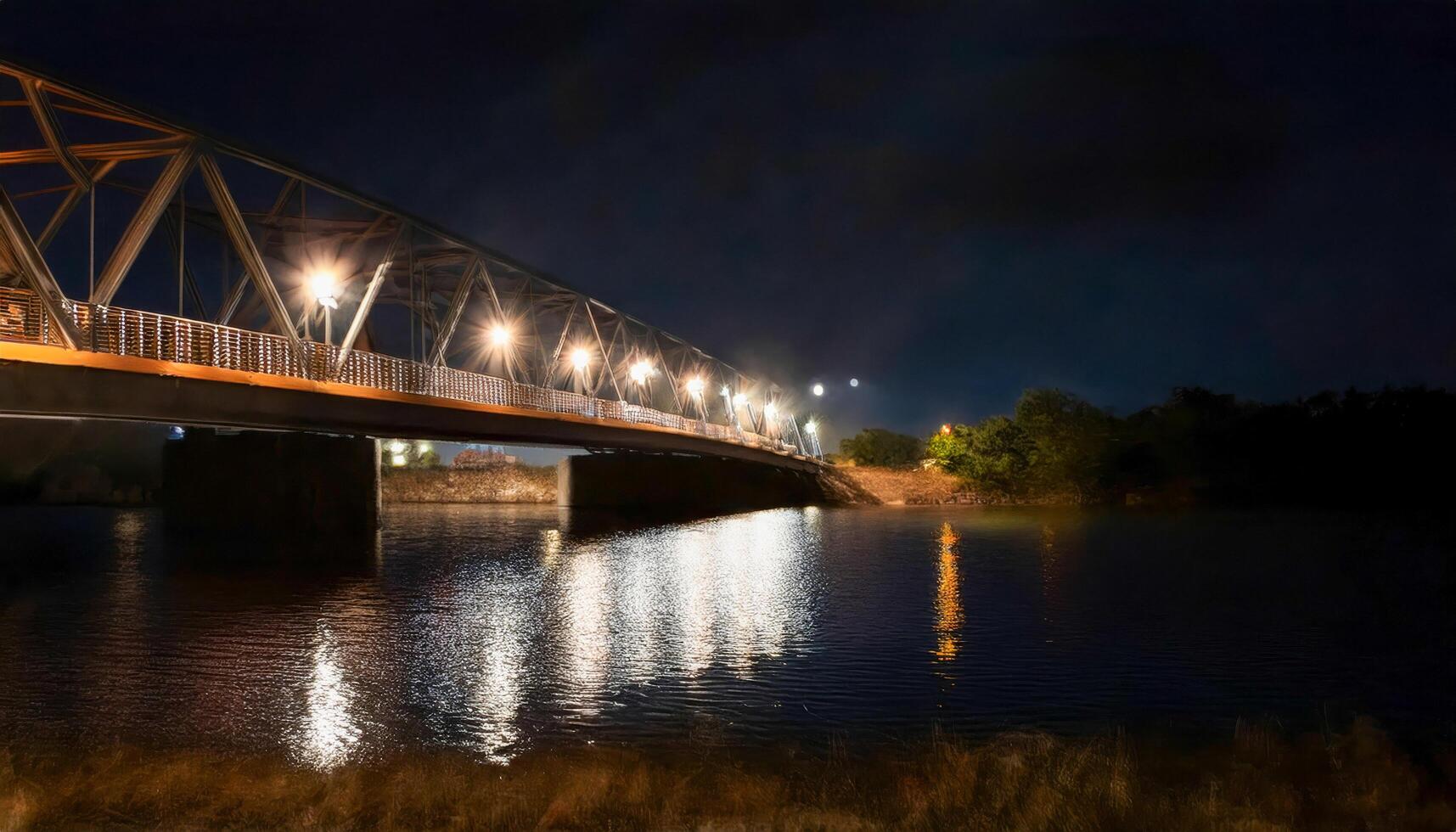 Bridge illuminated at night, lights reflecting on the dark river below. Truss structure spans the water. Tranquil and serene urban scene. Explore the night lights and reflections. photo