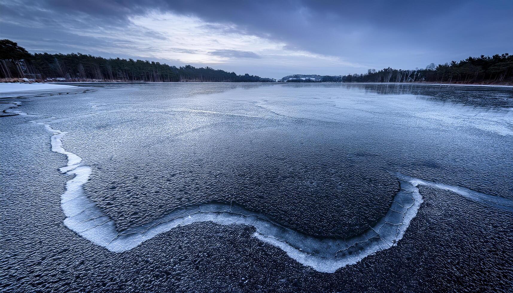 Wide view of a frozen lake with a prominent crack in the ice, under an overcast and moody sky. Winter landscape with trees lining the distant shore. Captures the cold stillness photo