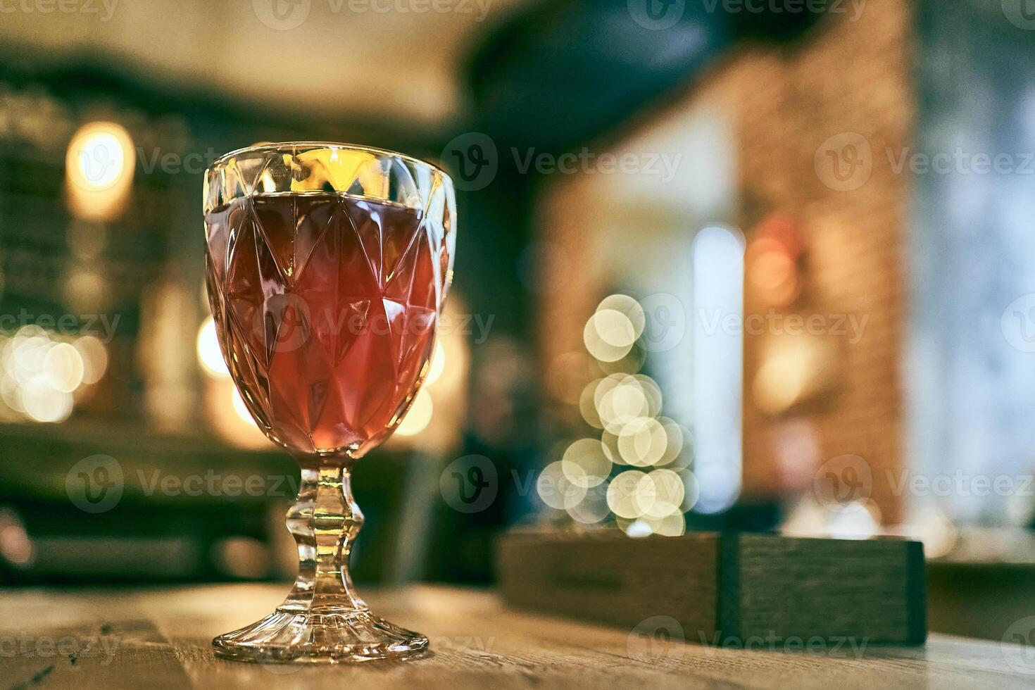 A filled wine glass with a traditional multifaceted shape, on a wooden bar counter with a blurred restaurant background with space to copy photo