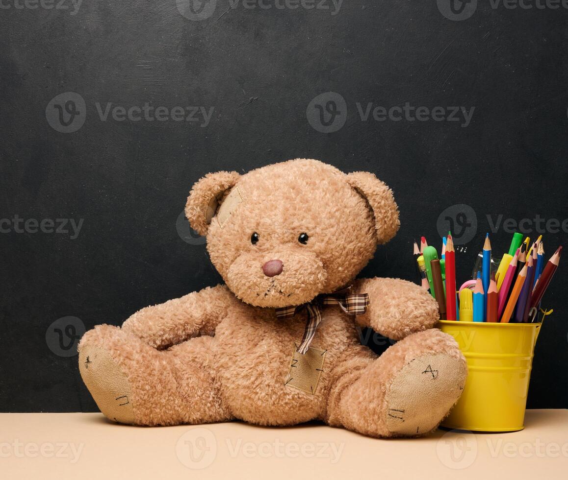 A brown teddy bear sits next to a yellow bucket filled with colorful pencils photo