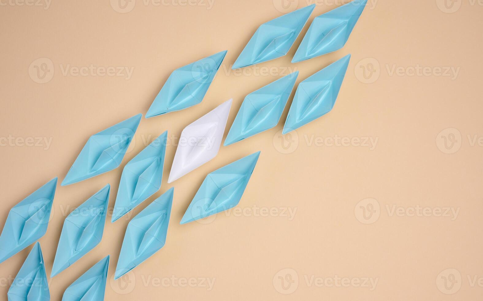 A white origami paper boat stands apart from a group of blue ones, symbolizing uniqueness or leadership, against a neutral background. photo