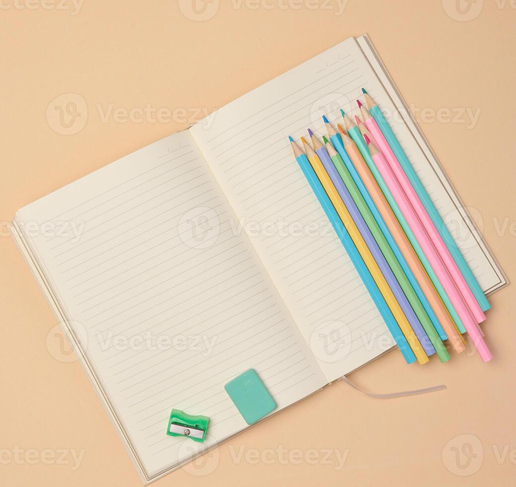 Top view of an open notebook with pastel-colored pencils, green sharpener, and turquoise eraser on a beige background. photo