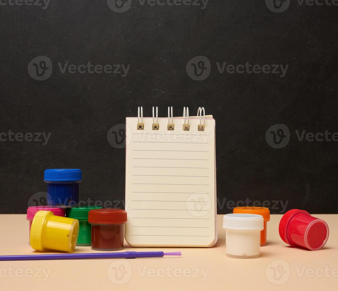 An open white spiral notebook surrounded by colorful paint cans and a brush photo
