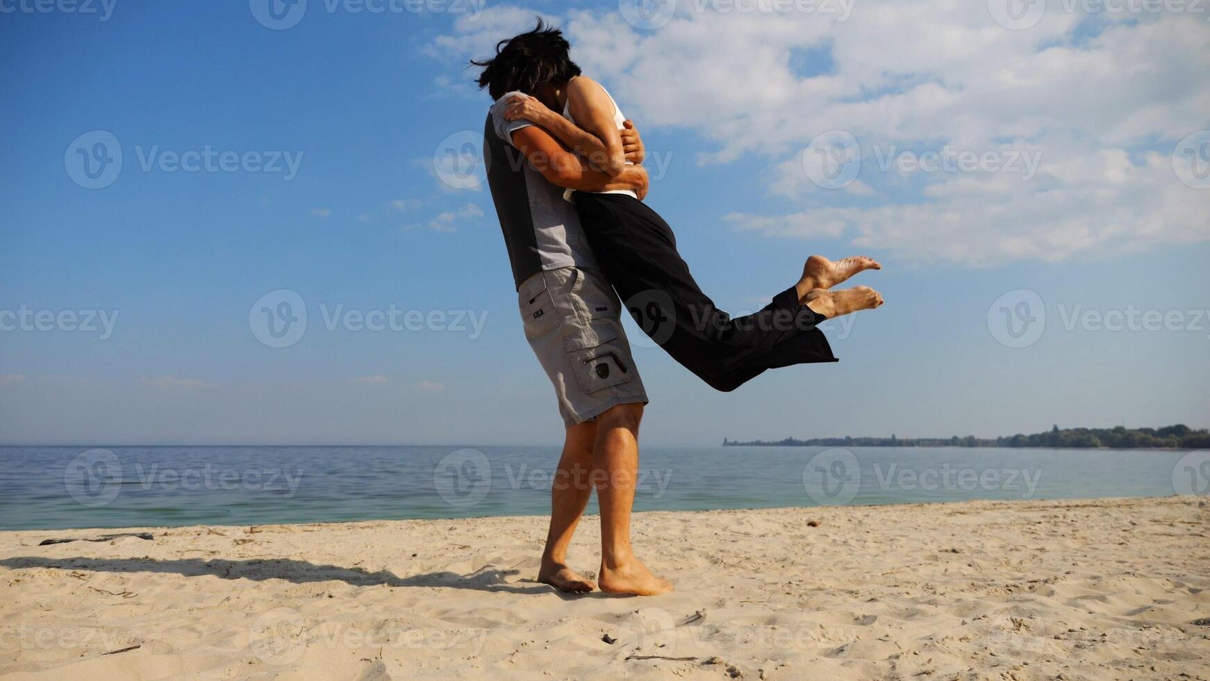 Happy couple having fun together on seashore at summer vacation. Man taking woman in air and turning around at coastline. Carefree lovers spending time at sandy beach. Concept of love. Slow motion photo