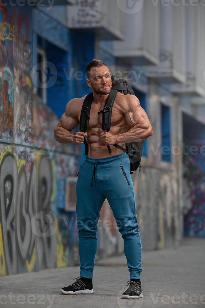 Muscular man poses confidently in urban setting with graffiti background during daylight photo