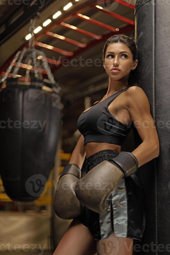 Strong female boxer practices in gym wearing gloves and sporty outfit while leaning against heavy bag during training session photo