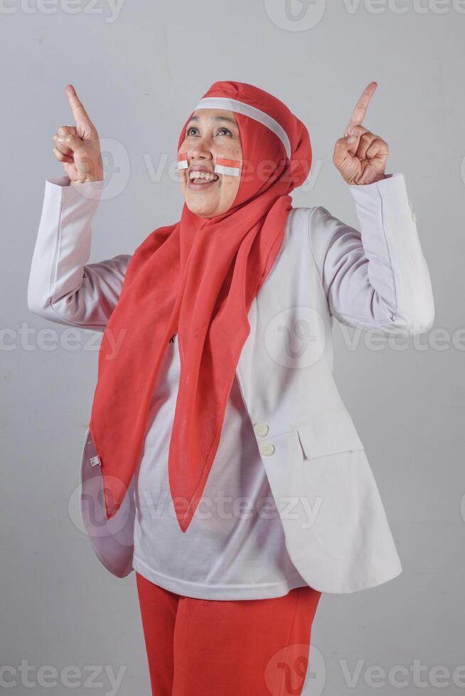 Happy Southeast Asian Muslim woman with patriotic face paint pointing up enthusiastically in celebration of national pride and cultural identity indoors photo