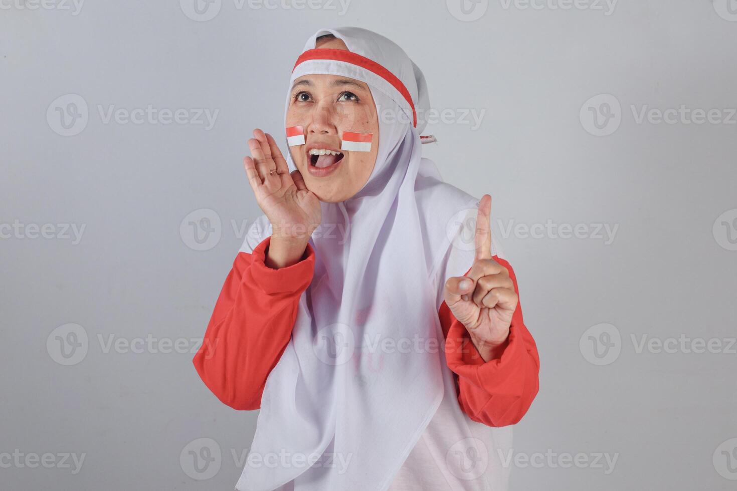 Muslim woman in red and white attire with Indonesian flag stickers on cheeks, cheerfully shouting and raising index finger to show enthusiasm and support during Independence Day celebration photo