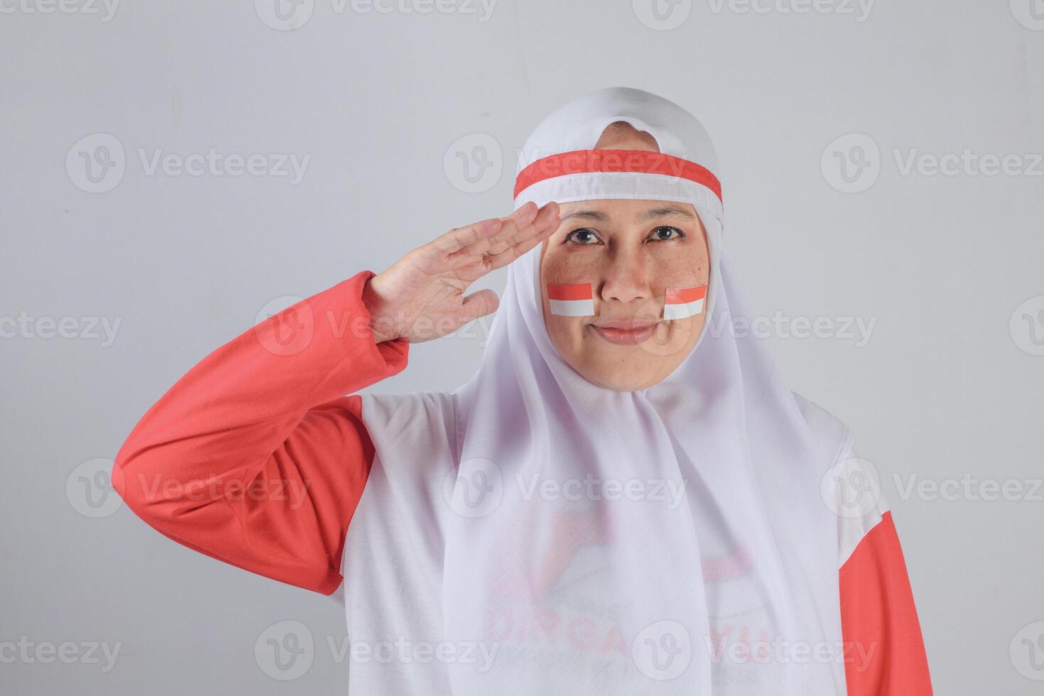 Indonesian muslim woman wearing red and white outfit saluting with patriotic expression and flag stickers on her cheeks, facing forward against plain background to honor Independence Day photo