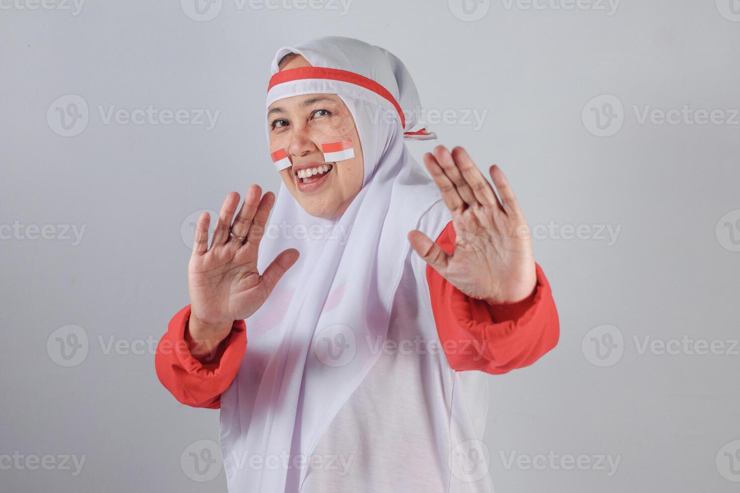 Indonesian woman in red and white hijab with flag face paint smiling and playfully holding hands up as if dancing or defending, radiating cheerful spirit during Independence Day celebration photo