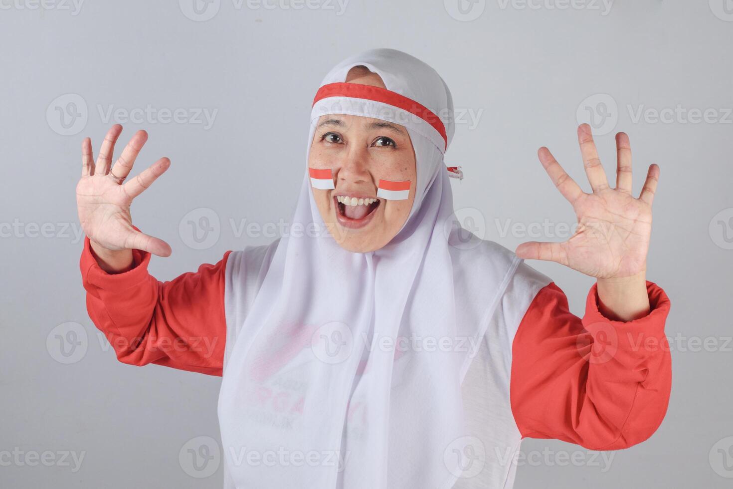 Excited Indonesian woman in red and white hijab costume celebrating Independence Day with painted flag on cheeks and enthusiastic open hands gesture showing patriotic spirit photo