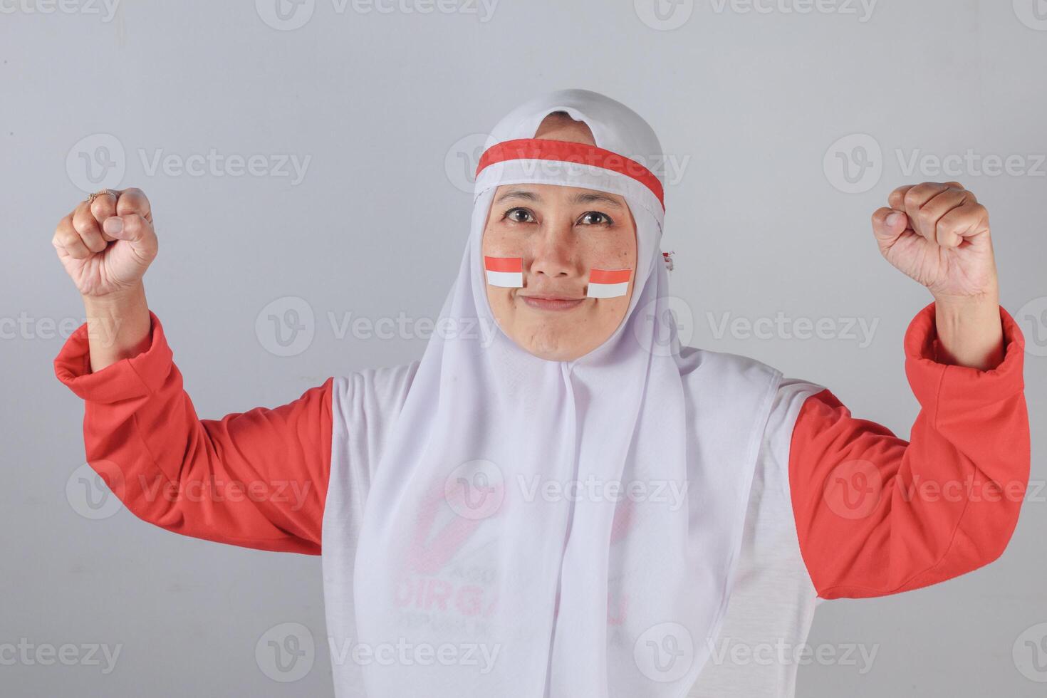 Confident Indonesian Muslim woman in white hijab and red clothing with patriotic face paint raising both fists in strong determined pose symbolizing national pride and empowerment photo