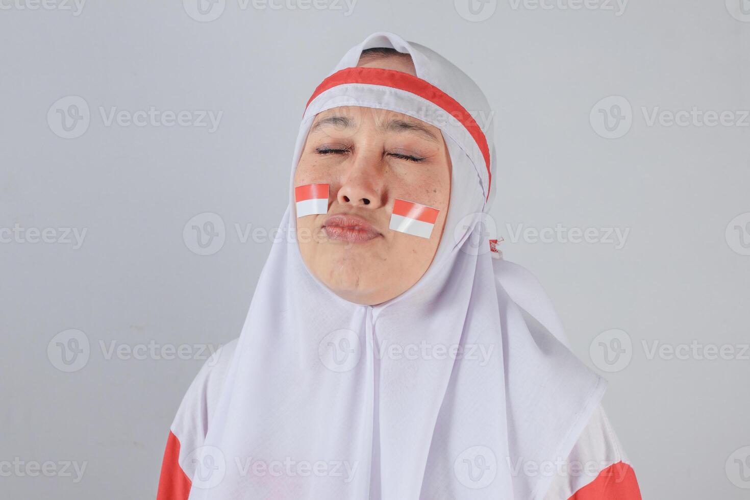 Indonesian woman in red and white hijab making funny pout expression with closed eyes and face paint of national flag symbolizing playful patriotic spirit during Independence Day photo