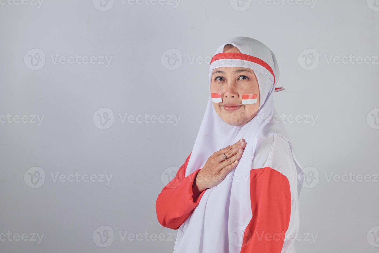 Muslim woman wearing red and white outfit with Indonesian flag stickers on cheeks, gently placing hand on chest and smiling with sincerity, symbolizing respect, gratitude, and national pride photo