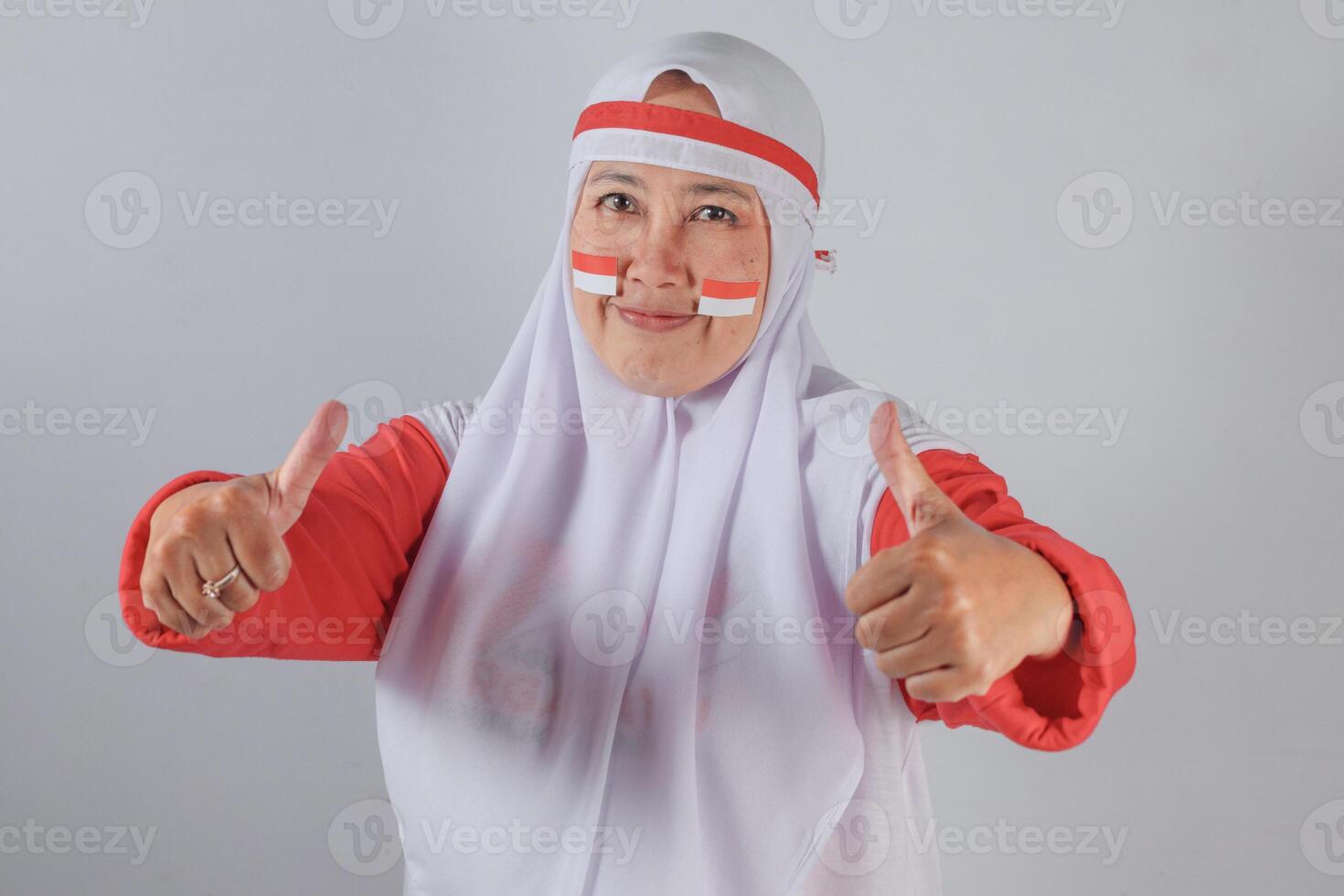 Indonesian Muslim woman in white hijab and red outfit with patriotic face paint showing double thumbs up and warm smile to celebrate Indonesian Independence Day with pride and positivity photo