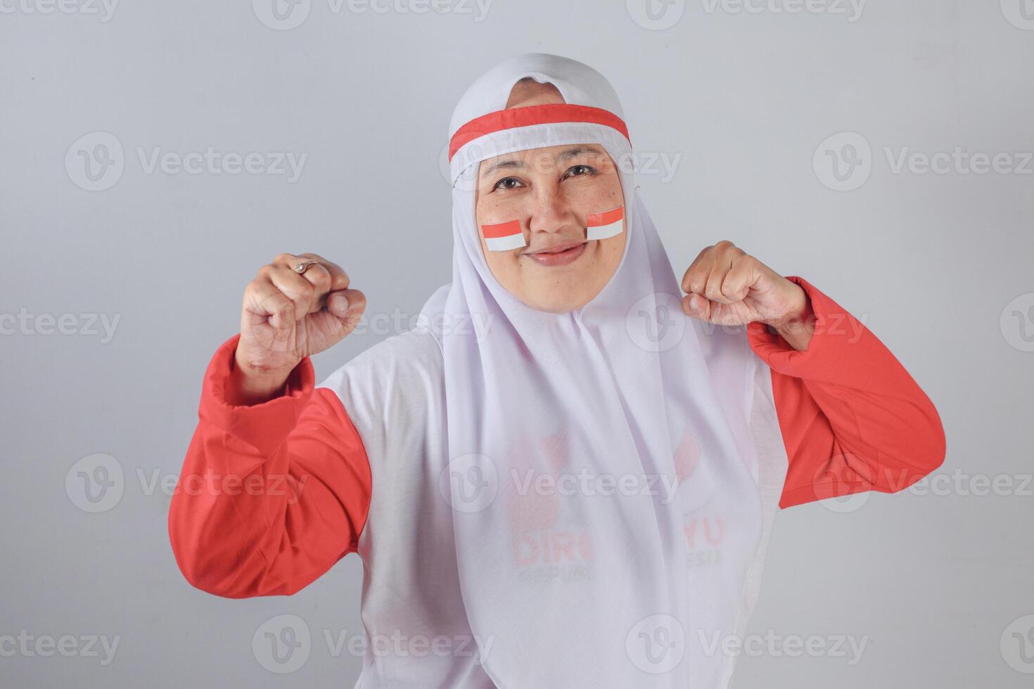 Smiling Indonesian Muslim woman in white hijab and red clothing with patriotic face paint raising both fists in a joyful and confident expression showing national pride and celebration photo