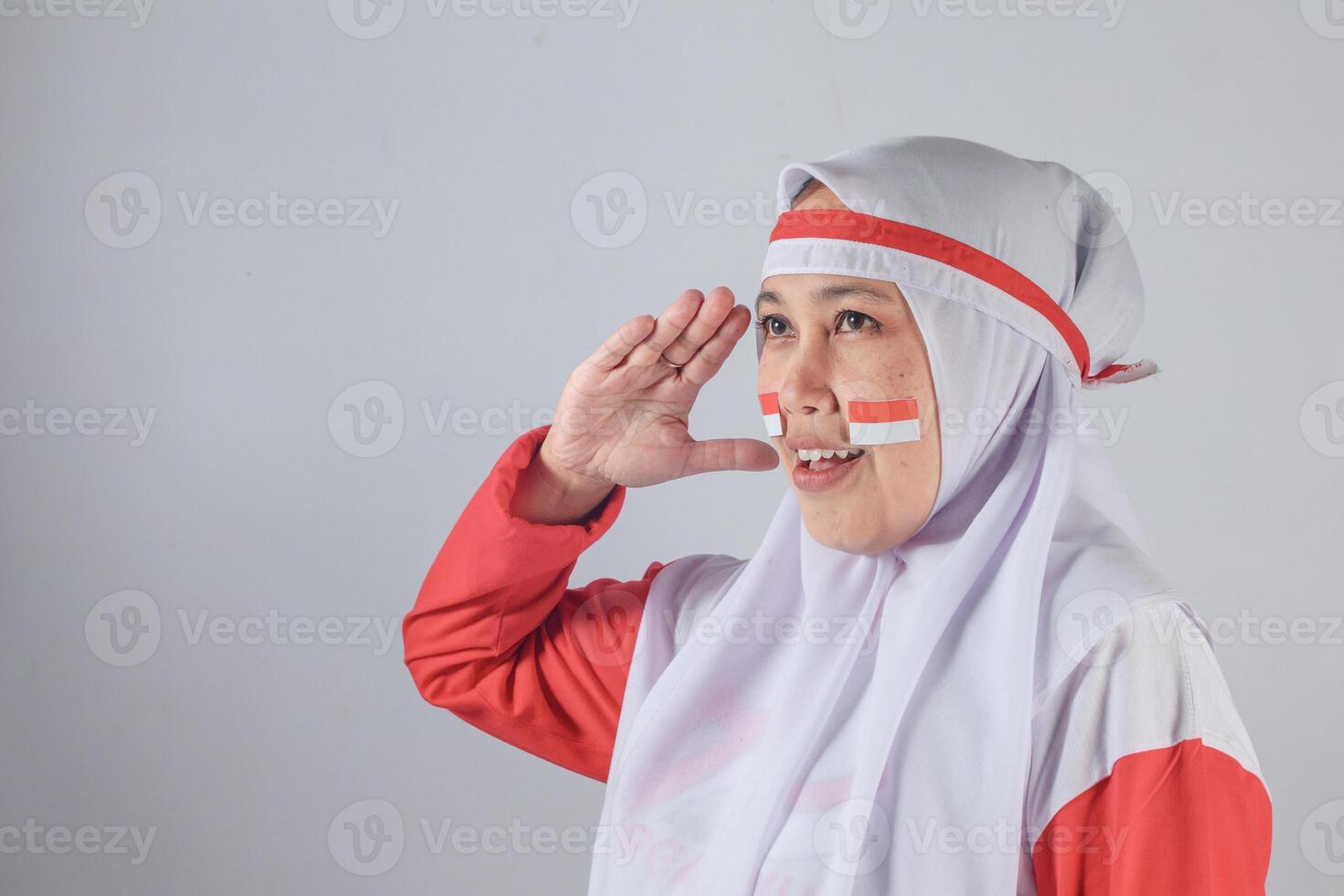 Muslim woman in red and white outfit saluting with Indonesian flag stickers on her cheeks, smiling proudly in front of a plain background, symbolizing national pride and patriotism on Independence Day photo