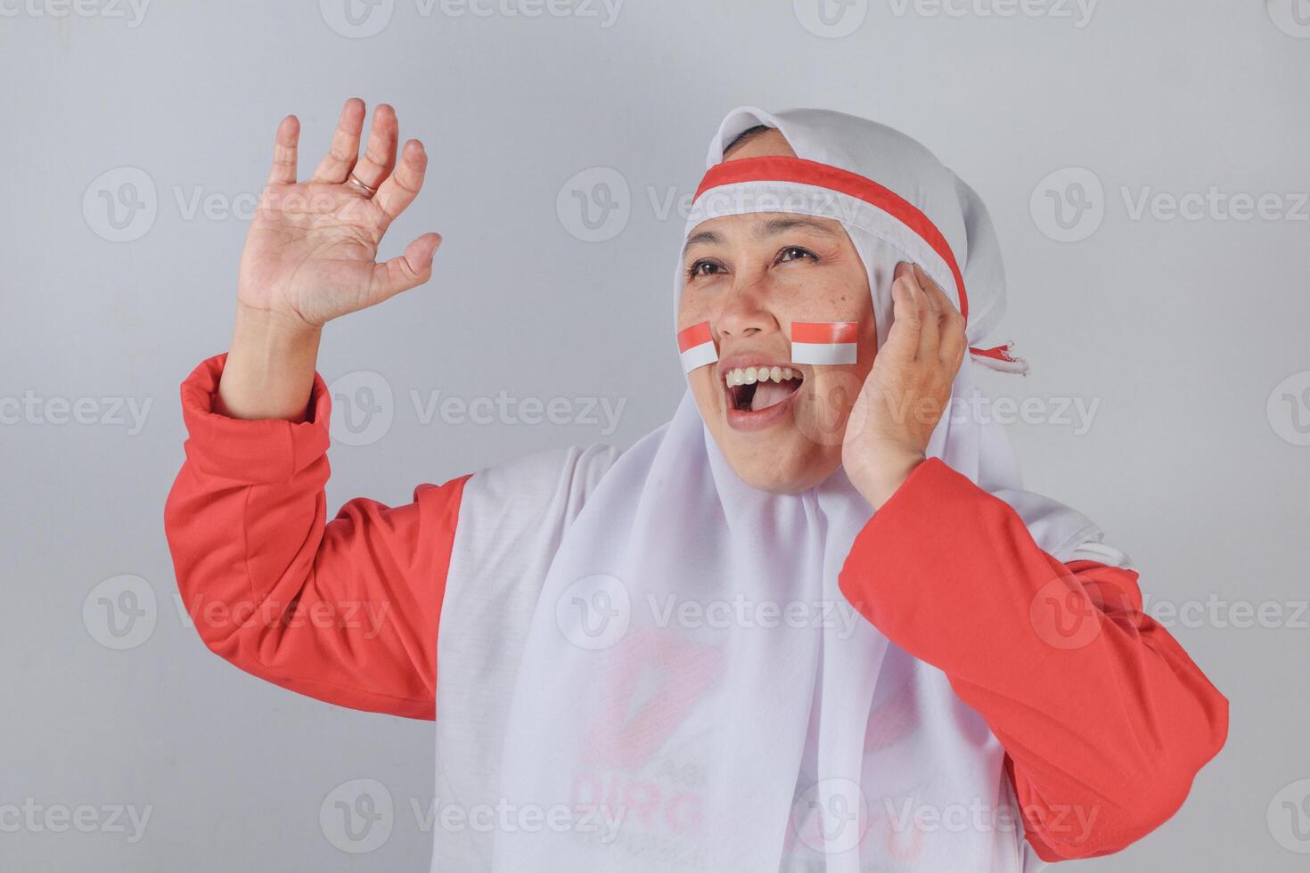 Playful Indonesian Muslim woman in white hijab and red outfit with flag face paint smiling joyfully while raising hands in a fun expressive pose during festive patriotic celebration photo