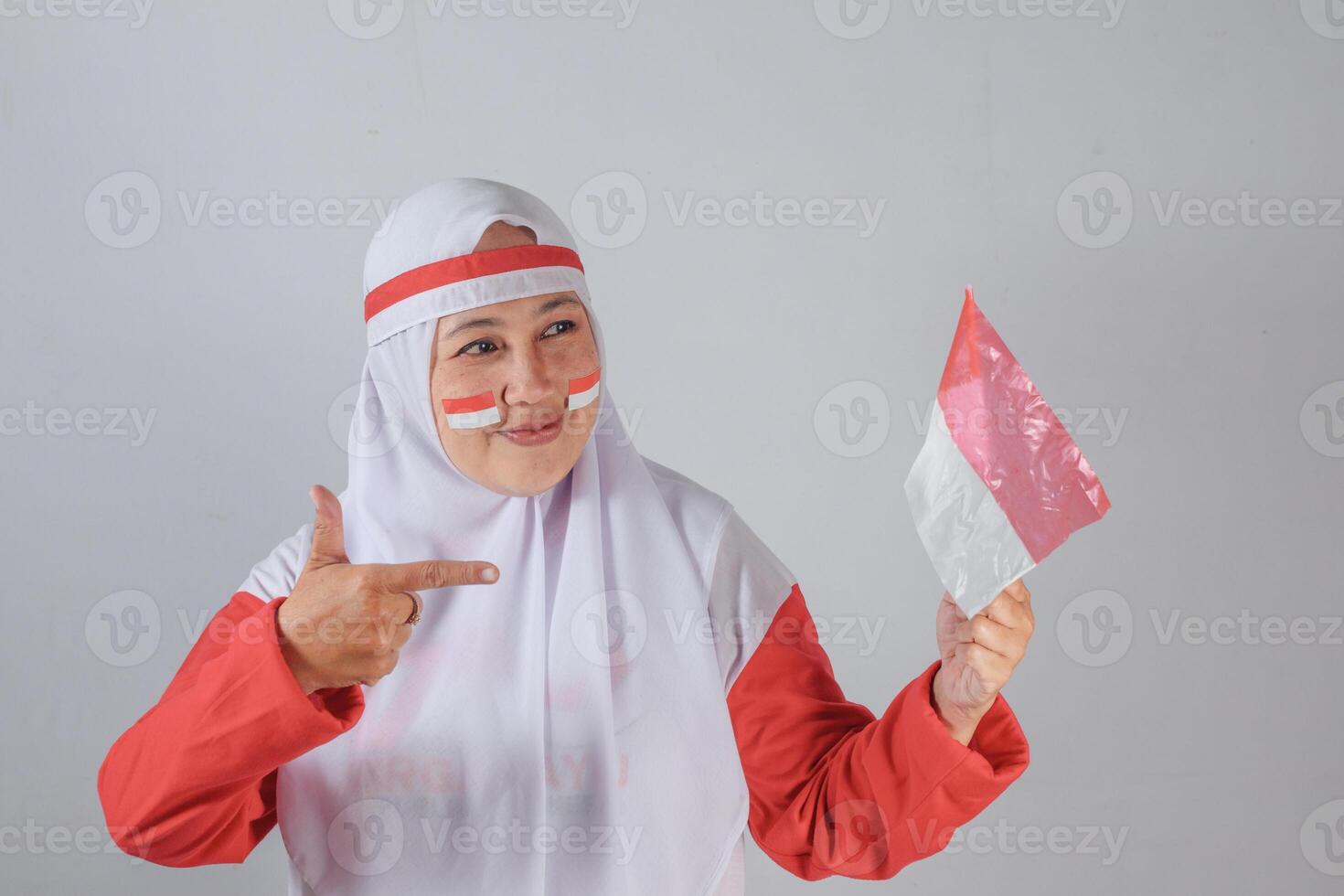 Smiling Southeast Asian Muslim woman in red and white hijab pointing proudly at small red and white plastic flag, celebrating national day with face paint and joyful expression photo