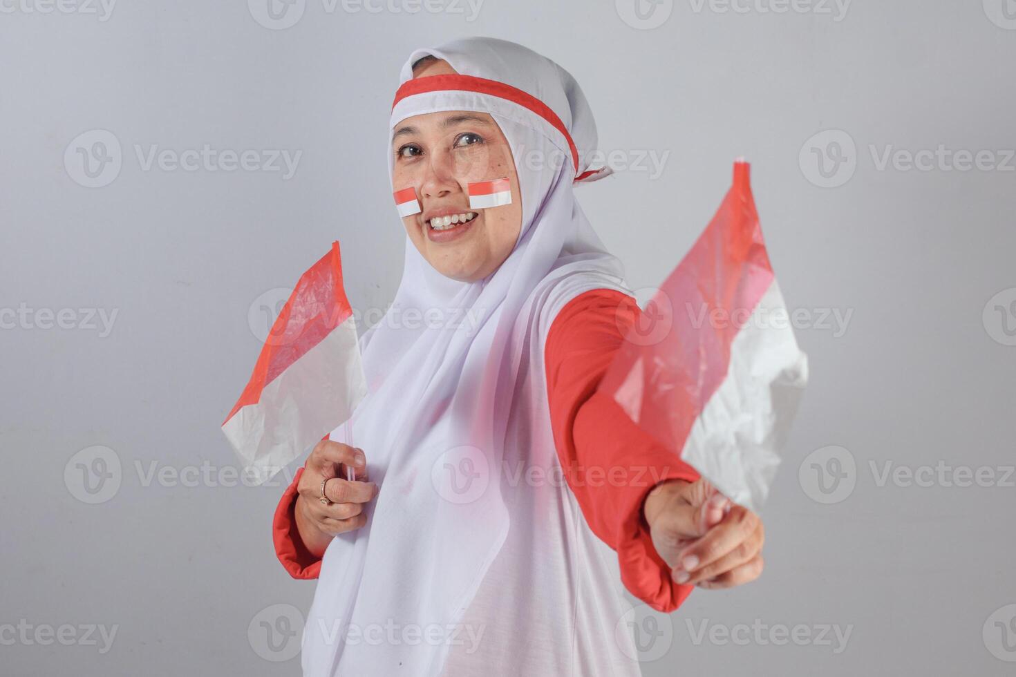 Smiling Muslim woman in hijab joyfully holding two Indonesian flags with face paint and festive expression during patriotic Independence Day celebration showing national pride and happiness photo
