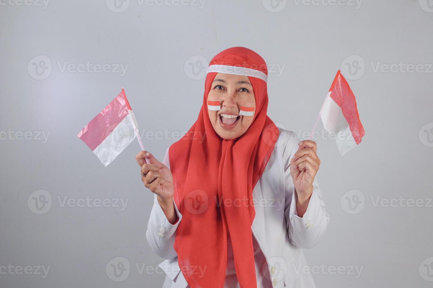 Excited Southeast Asian Muslim woman with face paint holding two national flags and expressing joyful energy during independence day celebration photo
