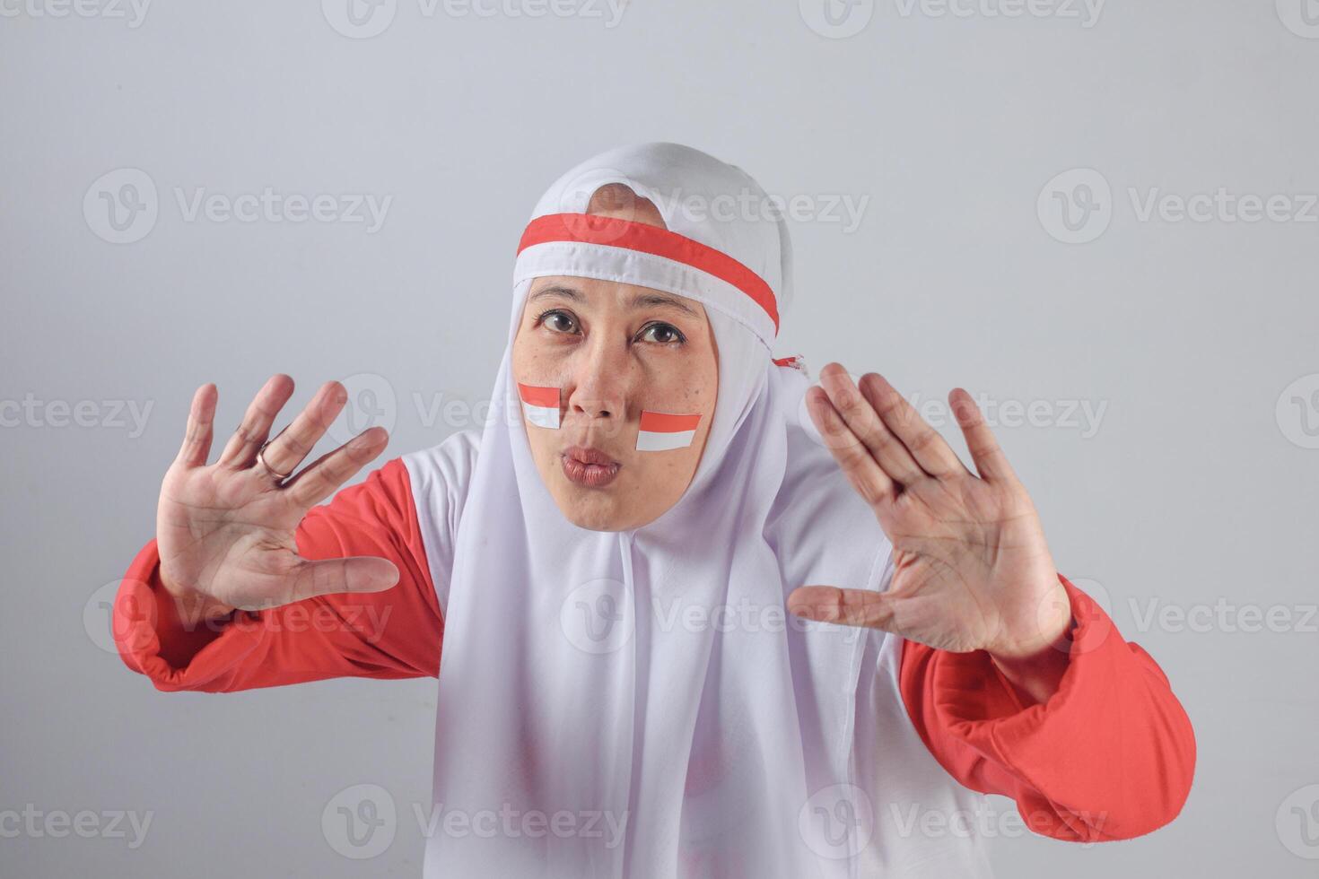 Indonesian woman in red and white hijab making puckered lips and playful hand gesture with flag face paint showing humorous and festive mood during Independence Day celebration photo