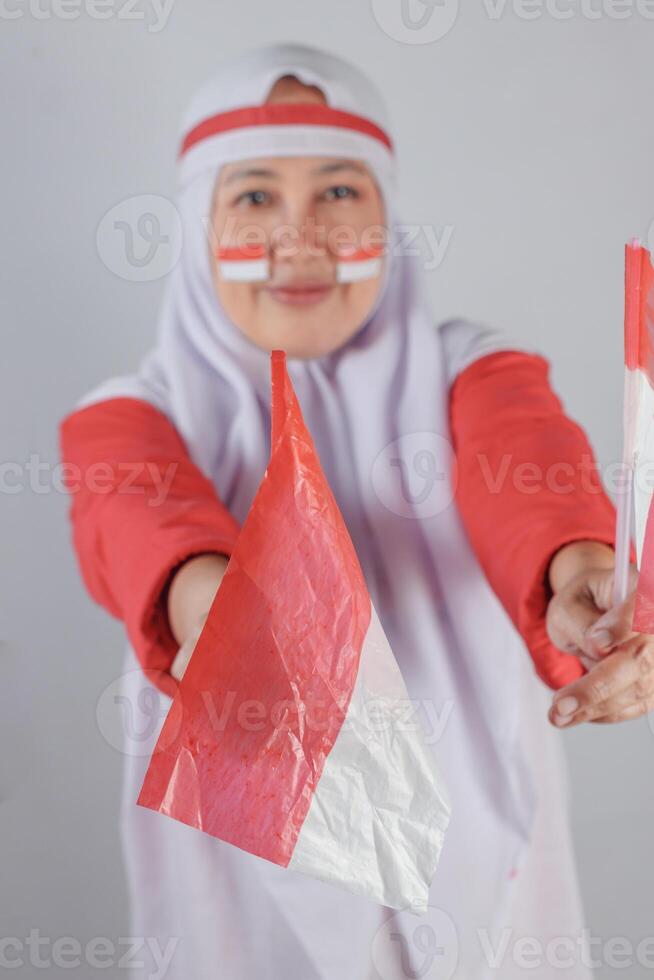 Muslim woman wearing red and white face paint and headband smiling proudly while holding small plastic Indonesian flags, celebrating Independence Day with patriotic spirit and festive emotion photo