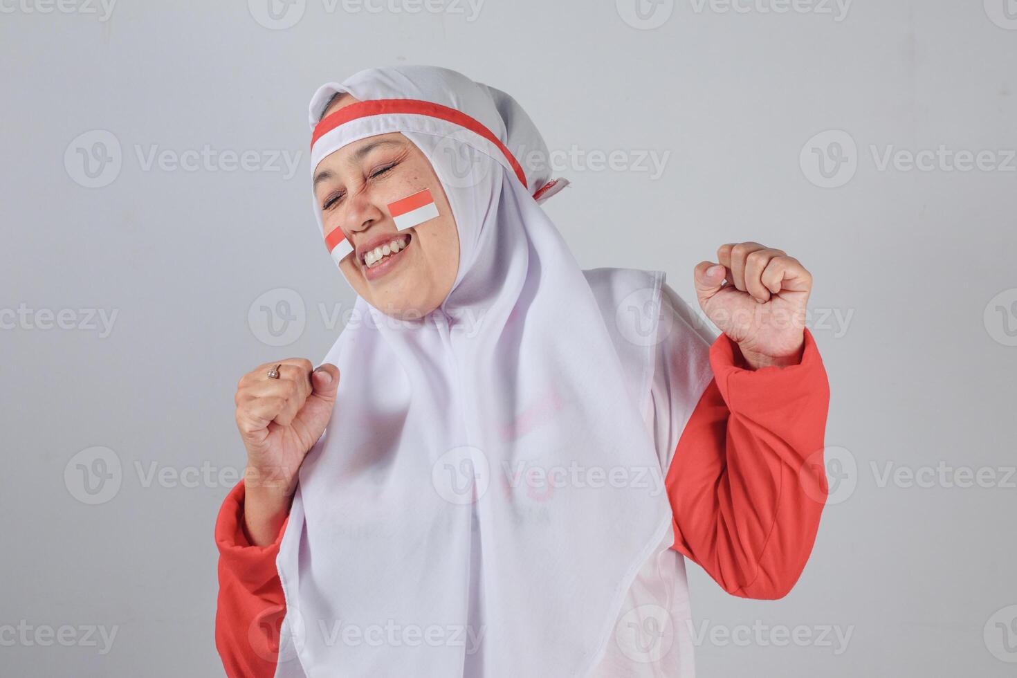Smiling Muslim woman in hijab with clenched fists celebrating national pride with Indonesian flag face paint, radiating joy, confidence, and cultural identity in expressive pose photo