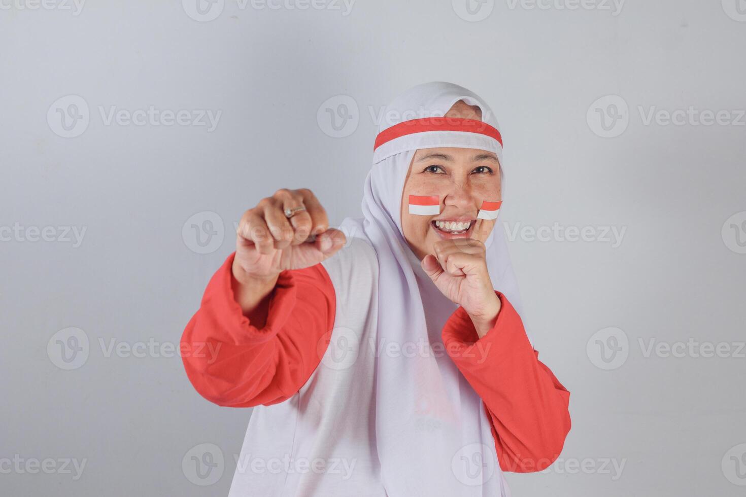 Energetic Indonesian woman in red and white hijab smiling brightly with fists raised in playful boxing pose and flag-painted cheeks during spirited Independence Day celebration photo