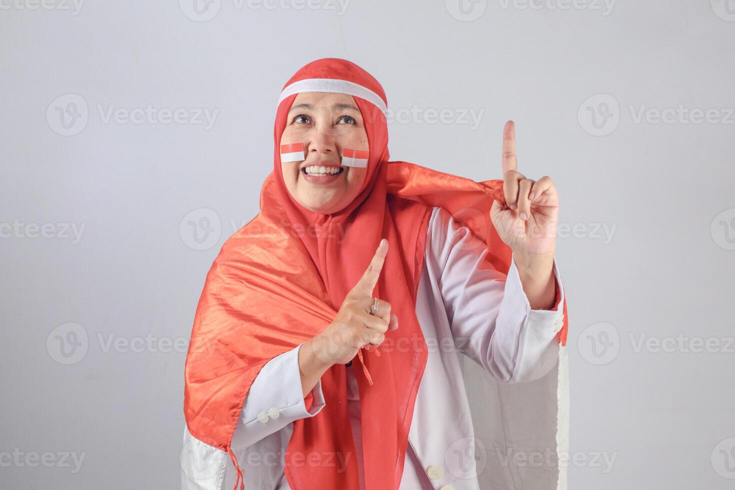 Southeast Asian Muslim woman wrapped in national flag smiling and pointing upwards with face paint celebrating independence and expressing joyful patriotism photo