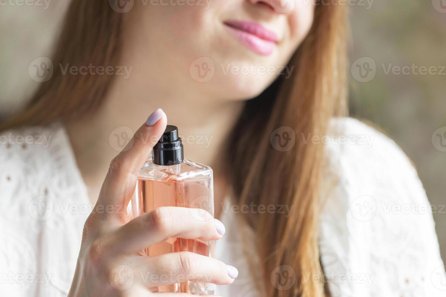 Beautiful young woman with bottle of perfume spraying it on her neck at home photo
