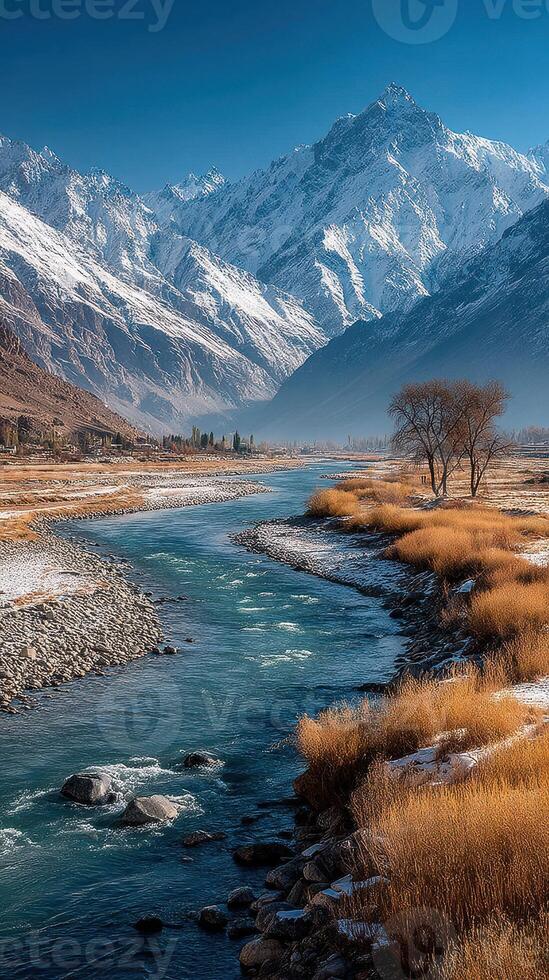Snow-capped mountains reflect in a serene river surrounded by golden grasses in a remote valley during daylight photo