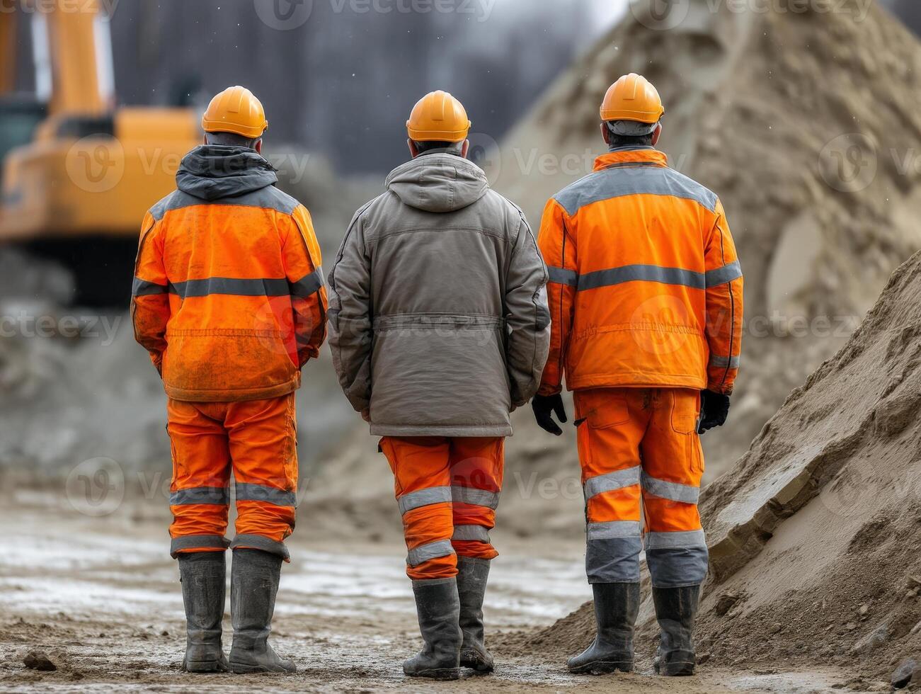 Three construction workers in orange safety gear and helmets stand on a sandy construction site, facing large equipment and mounds of sand under an overcast sky photo
