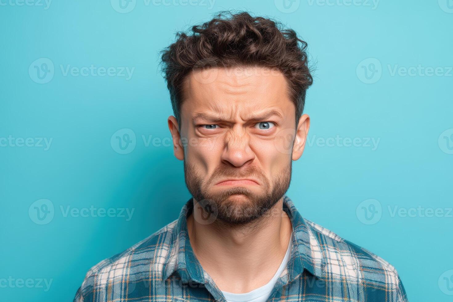 Young man with curly hair making a displeased facial expression, wearing a blue plaid shirt. His eyebrows are furrowed, and lips are pressed tightly together, conveying annoyance or frustration photo