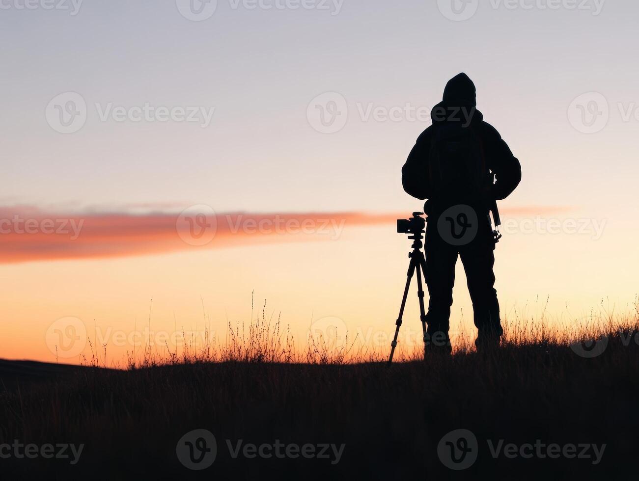 Silhouette of a person with a backpack and tripod, standing on grassy hill at golden hour. Sky displays gradient of orange to blue. Calm and peaceful outdoor scene suggestive of photography photo