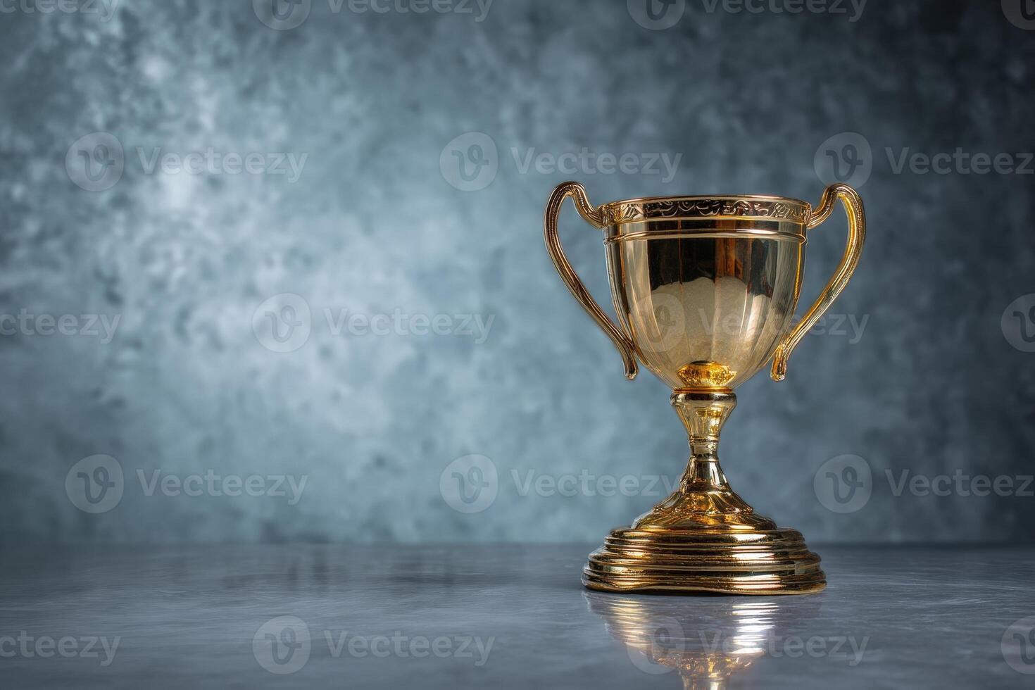 Golden trophy cup with ornate handles and detailed engraving, placed on a reflective marble surface with a smooth, soft-focus background. The cup's shine indicates high value photo